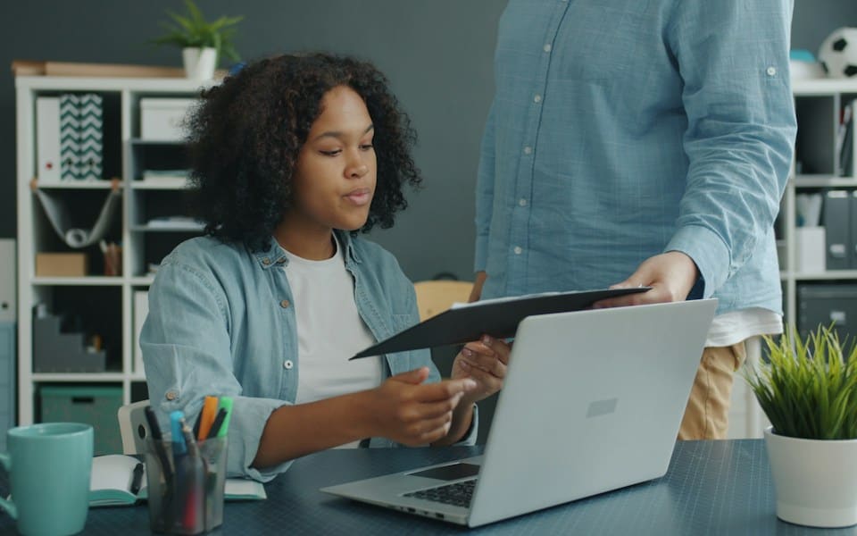A colleague handing a clipboard to a woman working at her laptop