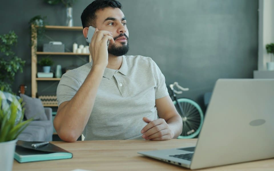 A man taking a phone call at his desk beside his laptop